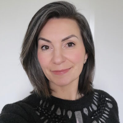 Een witte vrouw met zwart haar, met wat grijze stroken erdoor. Ze draagt een zwarte top en heeft een vriendelijke glimlach.
