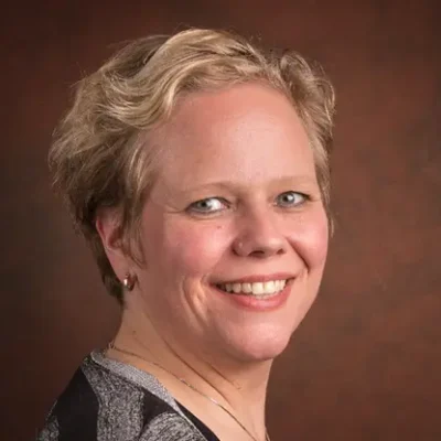 Vrouw met kort blond haar en oorbellen, lachend en zijwaarts gefotografeerd tegen een bruine achtergrond.