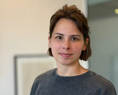 Vrouw met kort bruin haar in donkere trui, kijkend in de camera in een lichte binnenruimte.