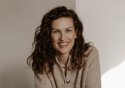 Lachende vrouw met krullend bruin haar met een lichtbruine trui aan, viir een beige achtergrond.