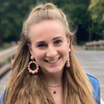 Vrouw met lang blond haar en grote oorbellen, lachend op een houten brug in een groene omgeving.