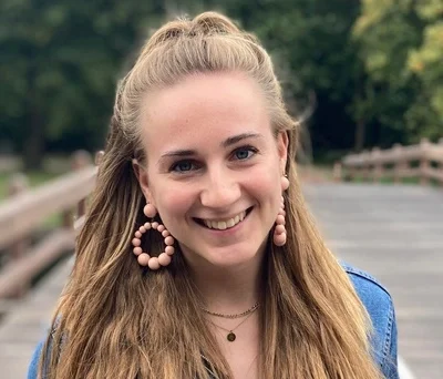 Vrouw met lang blond haar en grote oorbellen, lachend op een houten brug in een groene omgeving.
