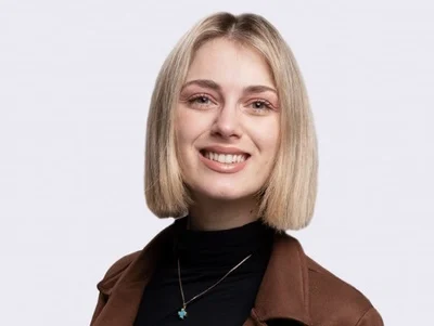Vrouw met blond bobkapsel en bruine blazer, lachend tegen een lichte achtergrond.