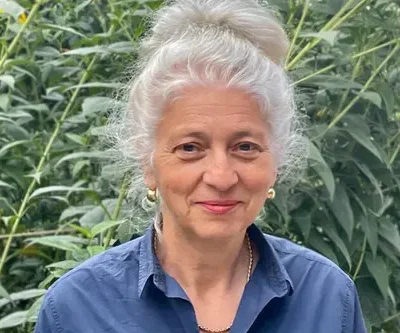 Portret van vrouw met opgestoken grijs haar, met een blauwe blouse aan en voor planten.