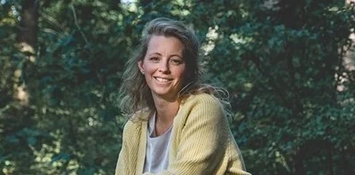 Lachende vrouw met halflang blond haar, gekleed in een geel vest en witte top, in een bosrijke omgeving.