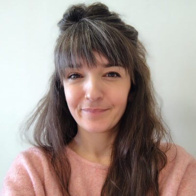 Foto van vrouw met bruin haar en grijze lokken. Vriendelijk lachend en in een roze trui.