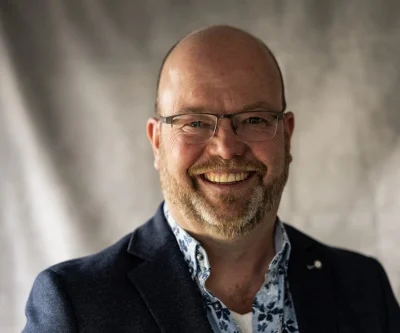 Portret van lachende man met een baard en bril, in een donkerblauw colbert en een licht overhemd, tegen een neutrale achtergrond.