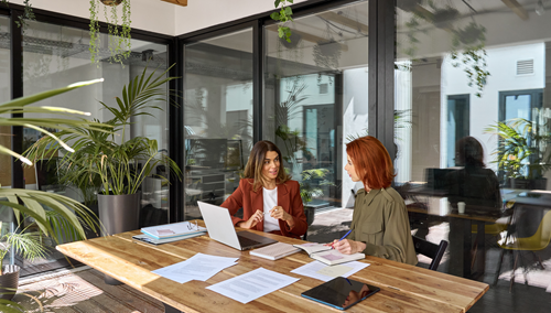 Twee vrouwen bespreken iets aan een tafel met daarop een laptop en papieren.